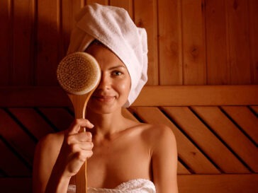 A young woman wearing a robe and relaxing in an infrared sauna while holding up a body brush to her face.