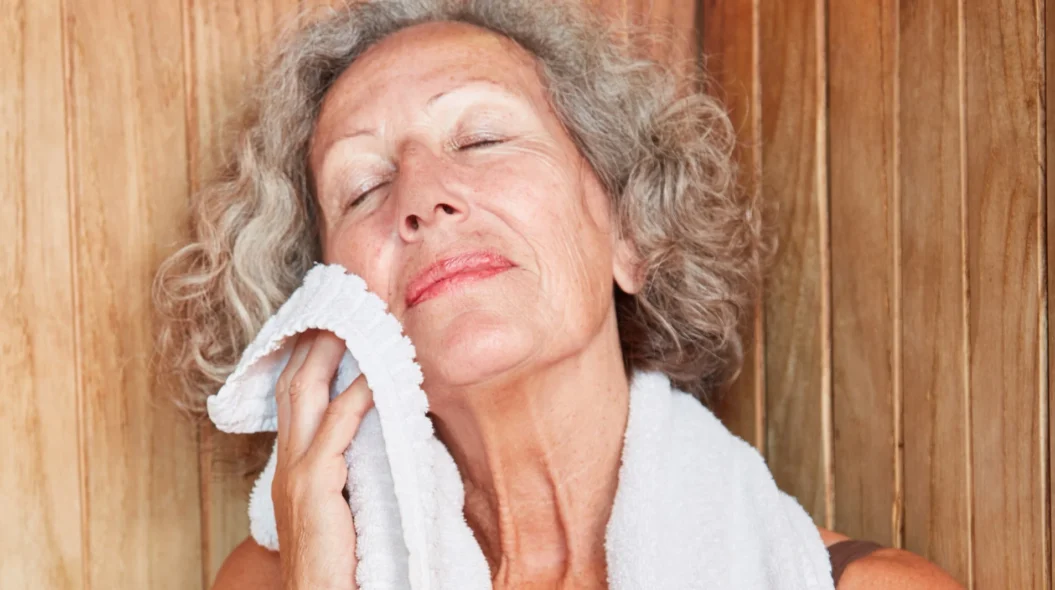An older woman relaxing in an infrared sauna while blotting her face with a soft towel.