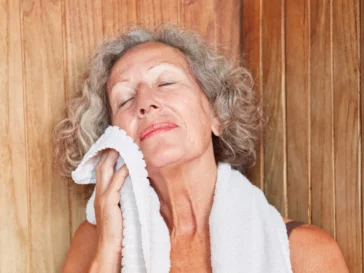 An older woman relaxing in an infrared sauna while blotting her face with a soft towel.